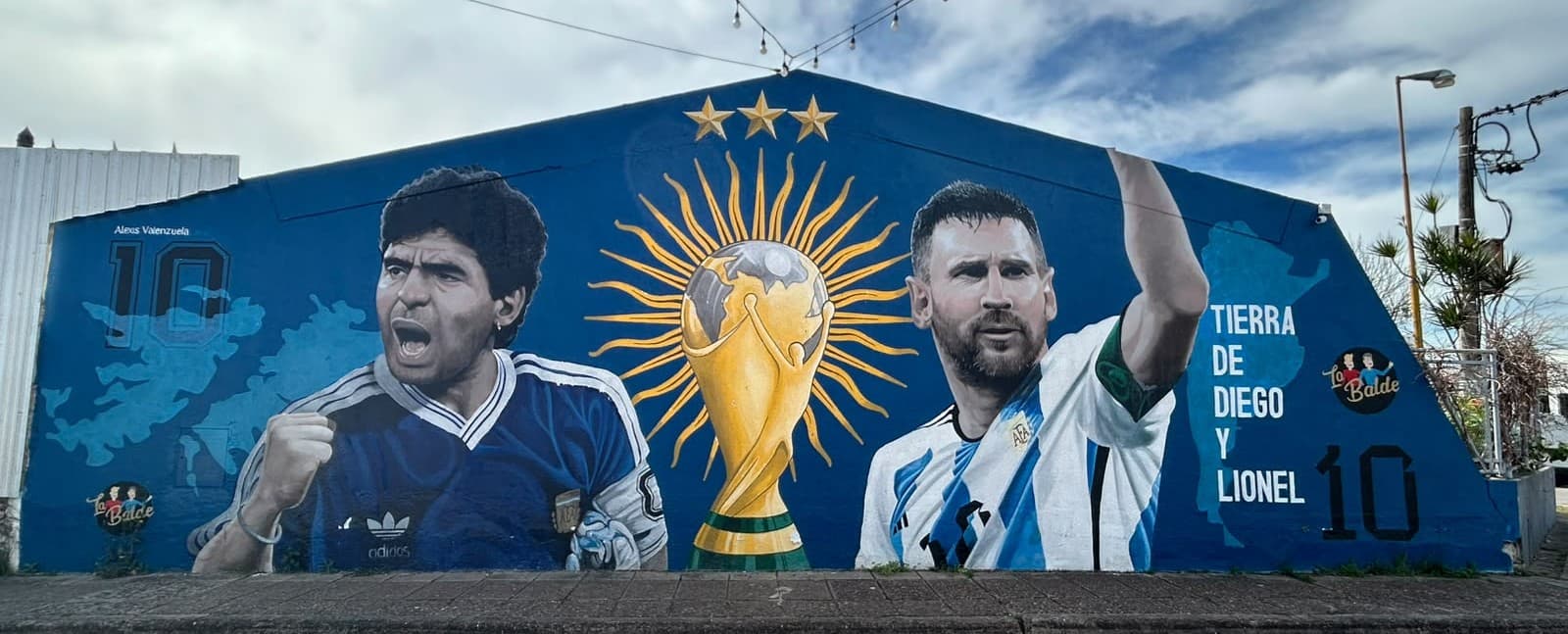 Mural callejero de Diego Maradona y Lionel Messi junto a la Copa del Mundo, sobre una pared azul.
